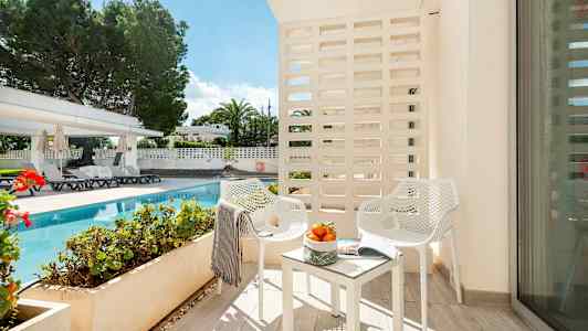 Billede af en balkon med to hvide stole og et bord med appelsiner, med udsigt til en pool og have. (AI-genereret tekst)