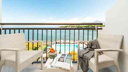 To hvide stole med et bord på en balkon, der giver udsigt over havet, en pool og landskabet. (AI-genereret tekst)