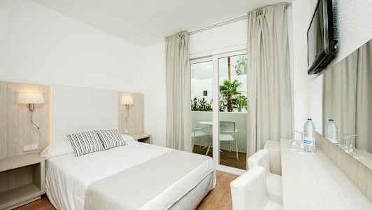 Lyst hotelværelse med dobbeltseng, skrivebord, stol og vindue med udsigt til en altan. (AI-genereret tekst)