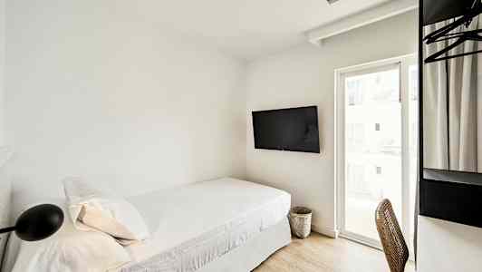 Et hotelværelse med en enkeltseng, vægmonteret TV og et skrivebord med stol ved siden af et vindue. (AI-genereret tekst)