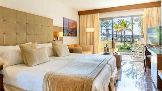 Hotelværelse med dobbeltseng, sofa, tv og store vinduer, der åbner ud til en balkon med udsigt over palmer og poolområde. (AI-genereret tekst)