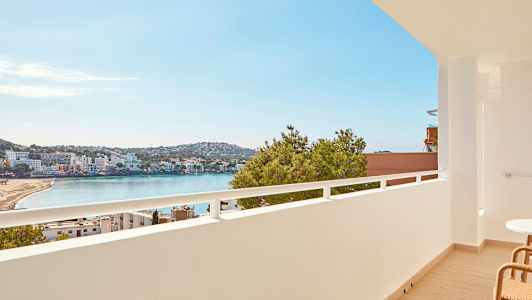 Udsigt fra en altan over en strand ved en bugt, med bygninger og grønt i baggrunden. (AI-genereret tekst)