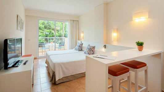 Et hotelværelse med to enkeltsenge, en lille siddegruppe og en altan med udsigt til grønne træer. (AI-genereret tekst)