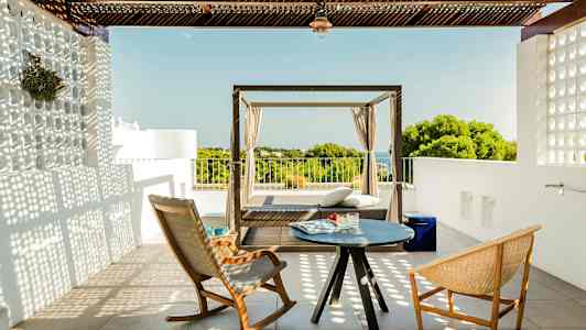 En terrasse med to stole og et lille bord samt en daybed med udsigt over grønt og hav. (AI-genereret tekst)