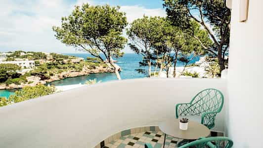 En balkon med to stole og et bord, udsigt over havet og landskab med fyrretræer. (AI-genereret tekst)