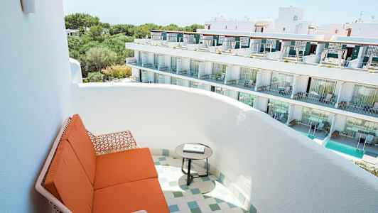 Billedet viser en altan med orange siddepladser og et rundt bord, med udsigt over en hvid bygning med flere etager og altaner. (AI-genereret tekst)