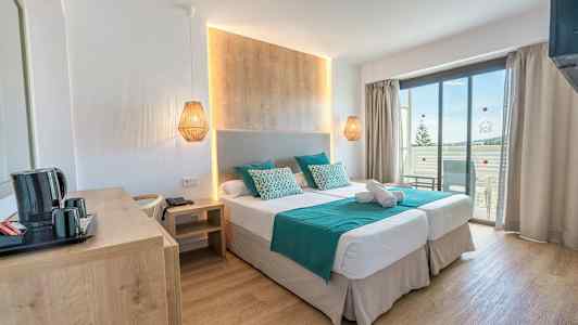 Et hotelværelse med to enkeltsenge, en træpanel bag sengen, turkis sengetøj og puder, samt en altan med udsigt. (AI-genereret tekst)