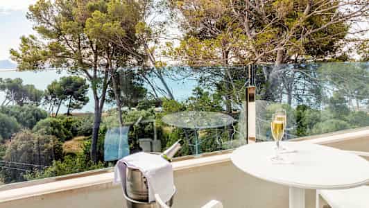 En balkon med udsigt over træer og havet. På balkonen står et hvidt bord med et glas champagne og en holder til en champagneflaske. (AI-genereret tekst)