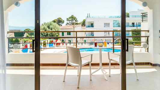 En balkon med to stole og et bord med drikkevarer og snacks. Udsigt over et poolområde og omkringliggende bygninger. (AI-genereret tekst)