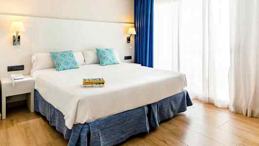 Et hotelværelse med en bred seng, hvid sengegavl, blå puder og gardiner samt et lyst trægulv. (AI-genereret tekst)