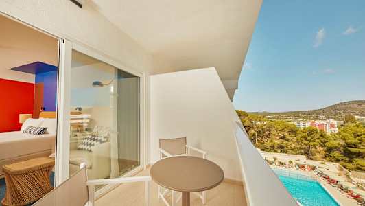 Udsigt fra en altan med bord og stole, med en pool og bygninger i baggrunden. (AI-genereret tekst)