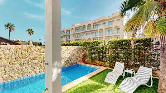 Billede af en udendørs pool omgivet af stenhegn og frodig have, med to hvide liggestole og et smukt hotel i baggrunden. (AI-genereret tekst)