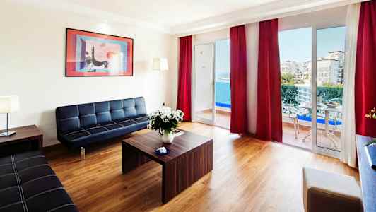 En moderne stue med udsigt til havet gennem store vinduer og balkon, møbleret med sorte sofaer og et træbord. (AI-genereret tekst)