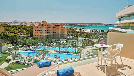 Udsigt fra en balkon over en pool og have med palmer, hotelbygninger og hav i baggrunden. (AI-genereret tekst)