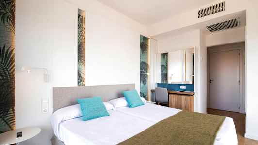 Et hotelværelse med to senge dekoreret med turkise puder og et grønt sengetæppe. (AI-genereret tekst)