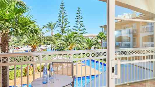 Et bord med flasker og glas på en altan med udsigt over palmer og en pool. (AI-genereret tekst)