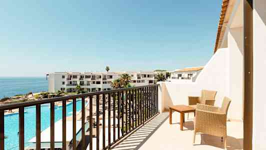 En altan med to flettede stole og et bord, udsigt over et hotelområde med udendørs pool og havet i baggrunden. (AI-genereret tekst)