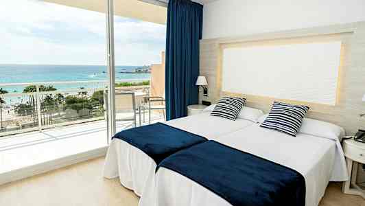 Et hotelværelse med to senge, blåhvide pyntepuder og blå sengetæpper samt en balkon med udsigt til havet. (AI-genereret tekst)