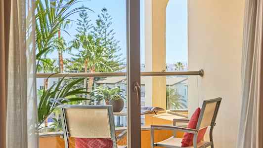 Udsigt gennem en balkon med to stole og et bord, omgivet af planter og bygninger. (AI-genereret tekst)