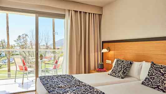 Billede af et hotelværelse med en dobbeltseng, et bord med stole på altanen og udsigt til palmer udenfor. (AI-genereret tekst)