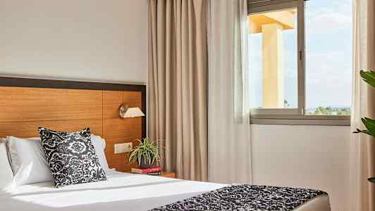 Hotelværelse med seng, natbord og et vindue, der lader naturligt lys komme ind. (AI-genereret tekst)