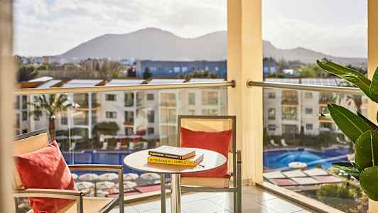 En udsigt fra en altan med et bord og to stole i forgrunden samt en pool nedenfor. Bjerge ses i baggrunden. (AI-genereret tekst)