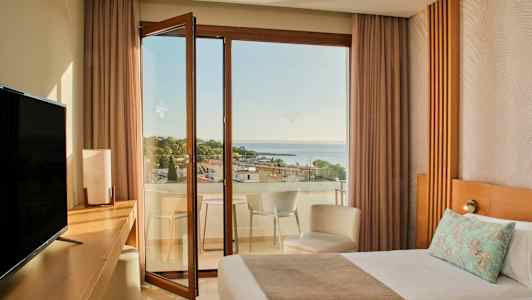 Et hotelværelse med en seng, skrivebord, TV og en åben altandør, der fører til en altan med udsigt over havet og træer. (AI-genereret tekst)