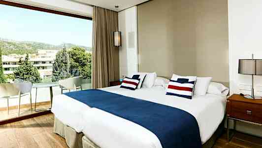 Et hotelværelse med en stor dobbeltseng, blå og hvide sengetæpper, samt et vindue med udsigt over beplantning og bygninger. (AI-genereret tekst)