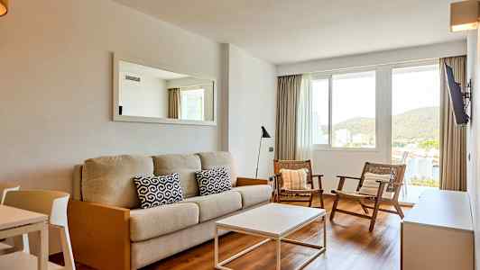 En stue med en beige sofa, to stole og et hvidt sofabord. Der er et vindue med udsigt til grønne omgivelser. (AI-genereret tekst)