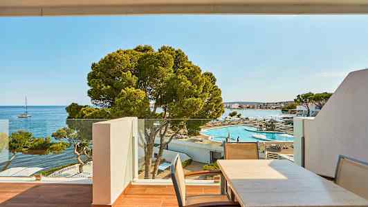 Udsigt over en udendørs terrasse med bord og stole mod et udendørs poolområde og havet med en sejlbåd i det fjerne. (AI-genereret tekst)