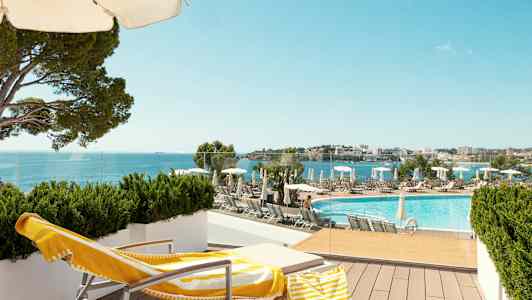 En liggestol med en gul-hvid håndklæde og strandtaske står på en terrasse med udsigt over en pool og havet. (AI-genereret tekst)