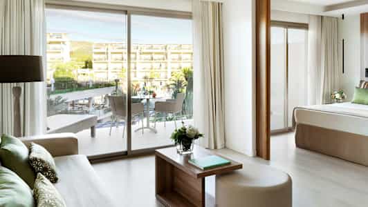 Udsigt fra et hotelværelse mod en balkon og et udendørsområde med pool og bygninger. (AI-genereret tekst)