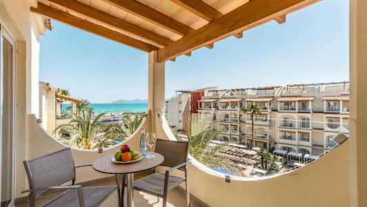 Balkon med to stole og et bord med frugtskål, havudsigt og hotelbygninger i baggrunden. (AI-genereret tekst)