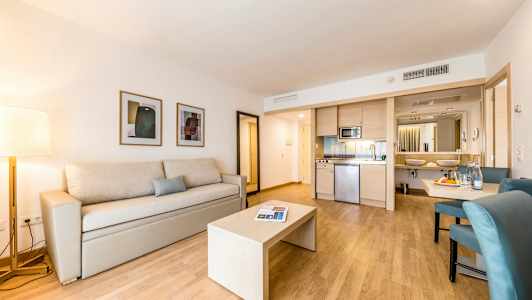 Et moderne hotelværelse med en beige sofa og bord på trægulv, tekøkken og badeværelse i baggrunden. (AI-genereret tekst)