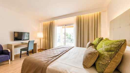 Et hotelværelse med en stor seng, grønne puder, et bord, en stol og et fjernsyn. Vinduer med gardiner lader naturligt lys komme ind. (AI-genereret tekst)