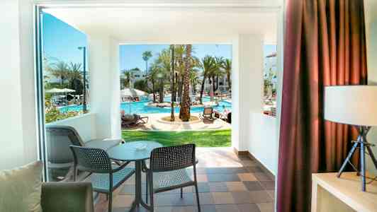 Udsigt fra et værelse med balkon mod et solrigt poolområde. (AI-genereret tekst)