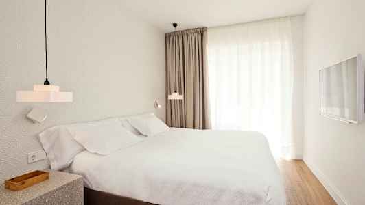 Værelse med en seng, hvide lagner og gardiner, samt vægmonteret TV. (AI-genereret tekst)