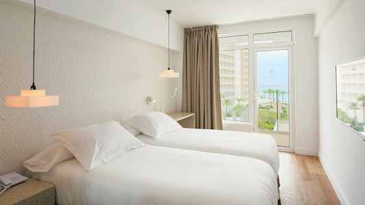 Et hotelværelse med to enkeltsenge, lyst interiør og en balkon med udsigt til havet. (AI-genereret tekst)