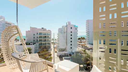 En altan med hvide stole og udsigt over moderne bygninger samt havet i baggrunden. (AI-genereret tekst)