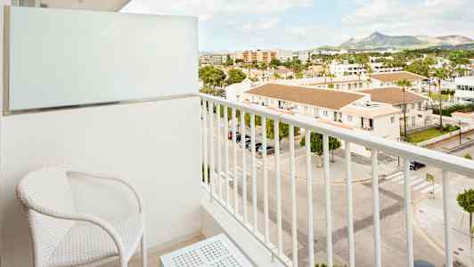 En balkon med to hvide stole og et bord, med udsigt over en gade og bygninger. (AI-genereret tekst)