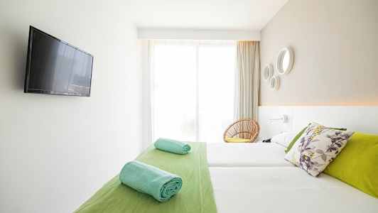 Et lyst hotelværelse med en dobbeltseng, farverige puder, sammenrullede håndklæder og et fladskærms-tv på væggen. (AI-genereret tekst)