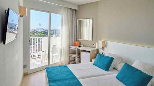 Et lyst hotelværelse med to senge, blå puder og sengetæpper samt en balkon med udsigt. (AI-genereret tekst)