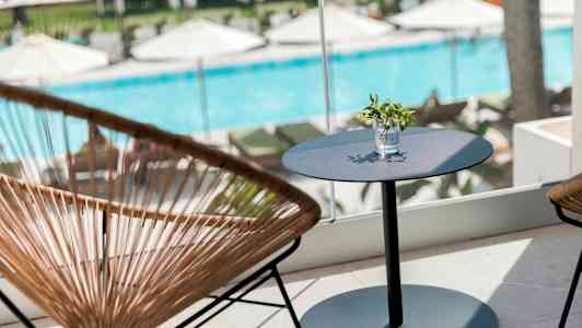 En siddegruppe med et bord og stole foran et vindue med udsigt til en pool og liggestole. (AI-genereret tekst)