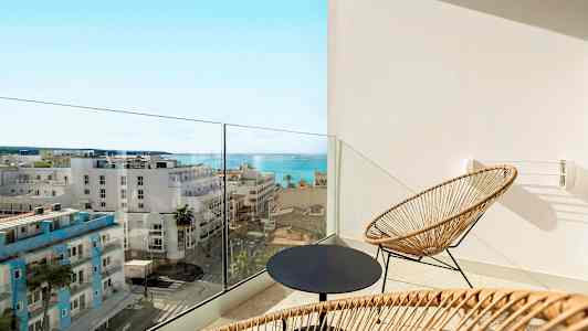 En altan med to stole i flet og et rundt bord, med udsigt over havet og byens bygninger. (AI-genereret tekst)