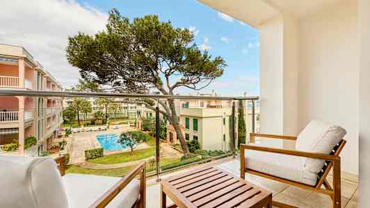 Udsigt fra en altan med siddemøbler, mod en gårdhave med swimmingpool og omkringliggende bygninger. (AI-genereret tekst)