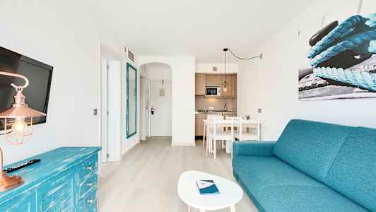 Et lyst hotelværelse med turkis tema, inklusive sofa, bord, tv og tekøkken. (AI-genereret tekst)