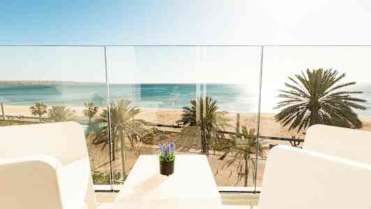 Udsigt fra en balkon mod havet og stranden med palmer. (AI-genereret tekst)