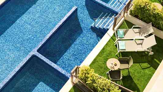 Billede af en swimmingpool med blå mosaik, to liggestole og et bord med stole på et grønt område. (AI-genereret tekst)