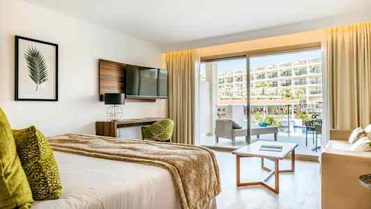 Et hotelværelse med en seng, sofagruppe og balkon med udsigt over poolområdet og hotellet. (AI-genereret tekst)