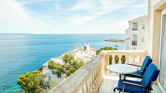 Udsigt fra en balkon med bord og stole over havet og omkringliggende bygninger. (AI-genereret tekst)